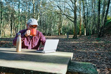 Olgun bir adam bardaktan içiyor, sıradan bir şekilde giyinip sahada dizüstü bilgisayarda çalışıyor.