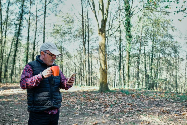 Olgun bir adam bardaktan içer, sıradan giyinir ve sahada cep telefonuna bakar.