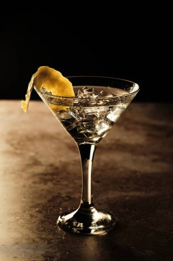 Sek bir martini kokteyli buzlu ve limon kabuklu berrak bir bardakta oturur. Arka plan karanlık, içkinin çıtır çıtır detaylarını vurguluyor..