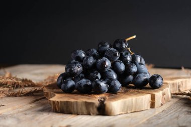 Dark grapes are placed on a wooden board. The background is dark, and the setting gives a simple and natural look to the grapes.