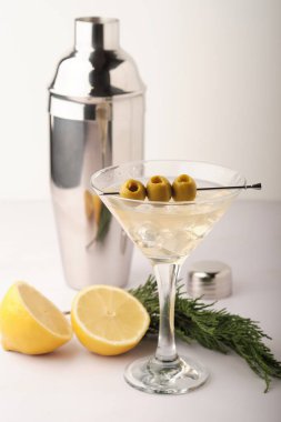 Üç zeytinli bir bardakta sek bir martini. Taze limon parçaları yakınlarda, bir titretici ve bazı otlarla birlikte, hepsi sade bir yüzeyde..