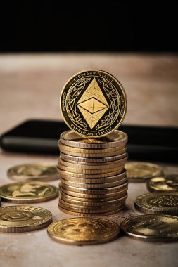 Bir masaya çoklu altın sikke konur. Üzerinde Ethereum tasarımı olan bir madeni para öne çıkıyor. Sikkeler ışığı yansıtır ve parlak bir görünüm yaratır..