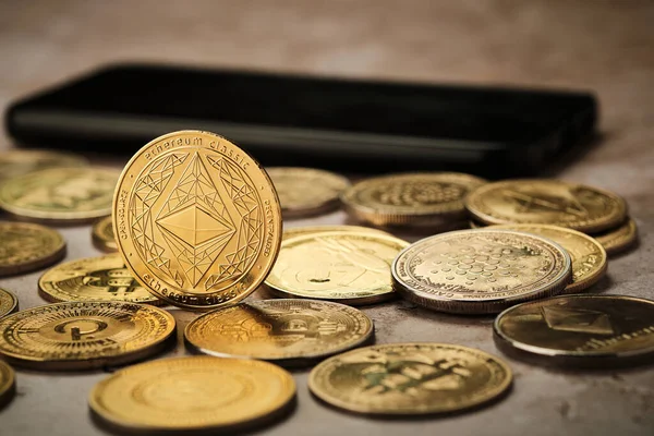 Birkaç altın kripto para birimi düz bir yüzeye yerleştirilmiştir. Bozuklukların yanında bir akıllı telefon görülebilir. Sahne klasik Ethereum madeni parasına odaklandığını gösteriyor..