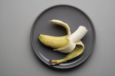 Bir muz, kabuğu kısmen alınmış gri bir tabağa yerleştirilir. Mutfak seti meyveyi ön plana çıkaran basit bir yemek düzenini gösteriyor..
