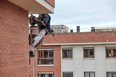 Bir işçi tuğla binaya tırmanıyor ve duvarı boyamak için bir silindir kullanıyor. Bu sahne, arka planda diğer binaların bulunduğu işlek bir şehir bölgesinde geçiyor..
