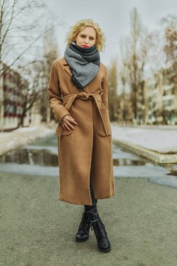 Kahverengi ceketli genç bir kadın kameraya bakıyor. Bulutlu bahar günü