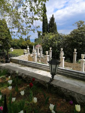 Ön planda nadir bulunan lale çeşitlerinden oluşan bir çiçek tarlası, eski taşların manzara tasarımı, dekoratif çalılar ve ağaçlar var..