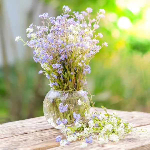 A bouquet of blue and white forget-me-nots in a glass vase on a wooden table in the garden. — Stock Photo, Image