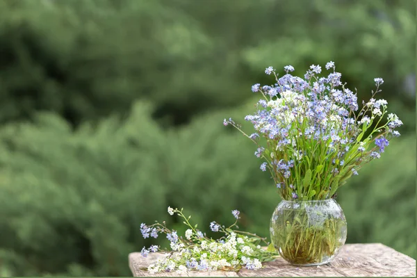 A bouquet of blue and white forget-me-nots in a glass vase on a wooden table in the garden — Stock Photo, Image