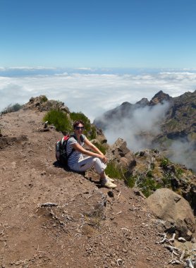 The girl with a backpack sits has a rest on a stony track highly in mountains, at the level of clouds