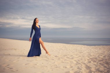 Kumlu bir plajda uzun elbiseli seksi bir manken. Kumaşlı gökyüzünün arka planında.