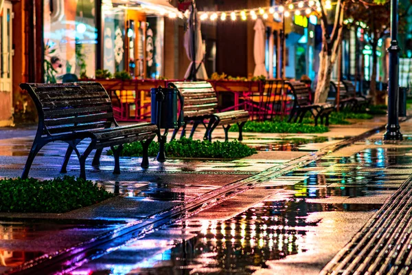 rain night cafe, autumn bench city. light outdoor lamp. street illumination after evening night rain. Night cityscape, colored lights reflected in wet asphalt