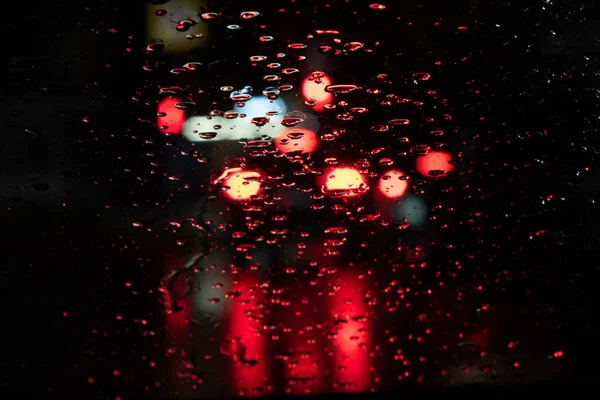 Rainy night traffic lights of big city, rain drops on car windshield glass, focus on raindrops. Rain background of a night blurred city. Rainy night weather. Outside the window is a blurred city