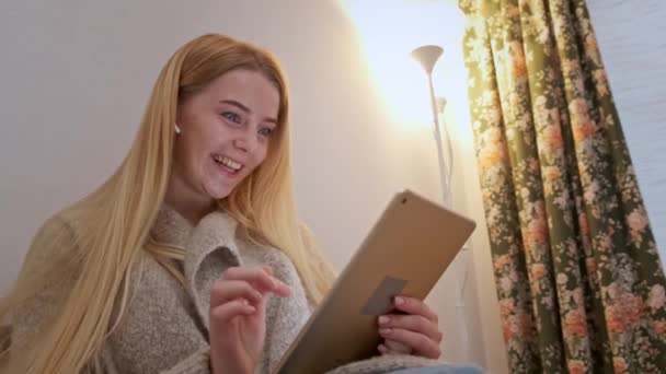 Joyeux sourire jeune femme assise sur le canapé, ami en appel vidéo à l'aide de la tablette — Video