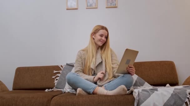 Onnellinen hymyilevä nuori nainen istuu sohvalla, ystävä videolla soittamalla tabletilla — kuvapankkivideo