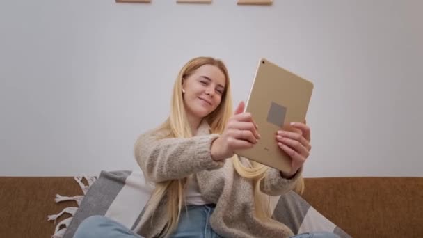 Joyeux sourire jeune femme assise sur le canapé, ami en appel vidéo à l'aide de la tablette — Video