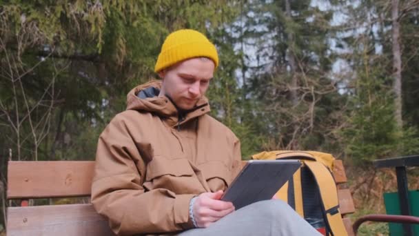 Ung man som har en videokall som sitter ute på bänken i skogen och pratar — Stockvideo