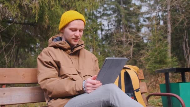 Молодий чоловік сидить на лавці в лісі, розмовляючи — стокове відео