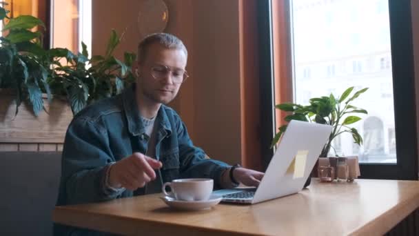 Young male freelancer working on a laptop sitting in the cafe having videocall — Stock Video