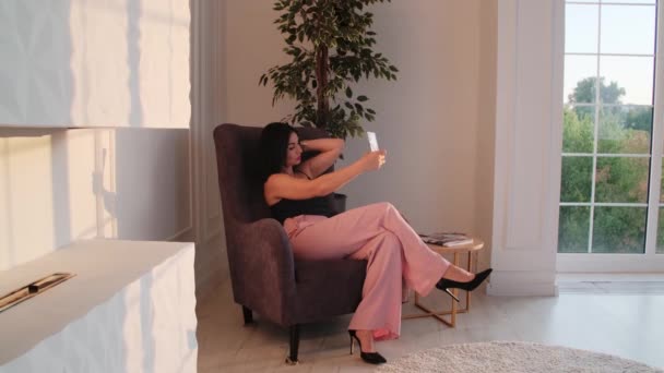 Hermosa mujer morena sonriente sentada en el sillón marrón usando su teléfono, hablando. — Vídeos de Stock