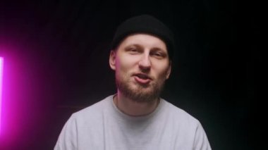 Siyah şapkalı genç bir adam, siyah bir arka planda el hareketleri yaparken pozitif duygularını gösteriyor..