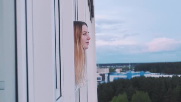 Κοντινό πλάνο ενός κοριτσιού που κοιτάζει έξω από το παράθυρο του μπαλκονιού απολαμβάνοντας τη θέα στο ηλιοβασίλεμα και κοιτάζοντας κάτω στο έδαφος. — Αρχείο Βίντεο