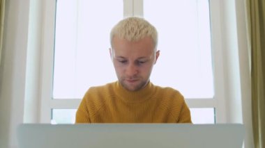 Genç Avrupalı bir erkek, evinde, masada bir fincan çay içerken dizüstü bilgisayarını kullanıyor..