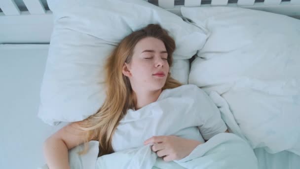 Belle jeune fille européenne se réveille le matin et regarde la fenêtre tout en restant au lit bâillant. Intérieur moderne blanc. — Video