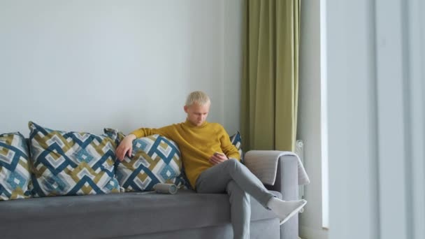 Giovane maschio attraente seduto su un divano grigio in un bellissimo appartamento leggero utilizzando il suo multitasking telefono. — Video Stock