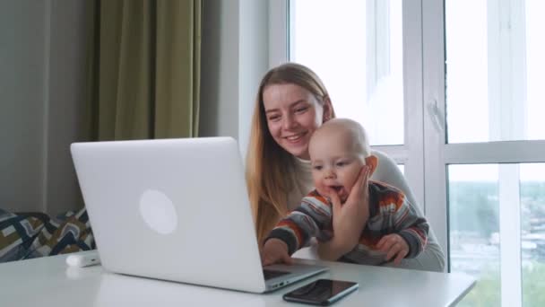 Young happy mother and her little baby son are having a videocall with the family. Sitting at the table in front of the big window. Smiling and laughing. — Stock Video