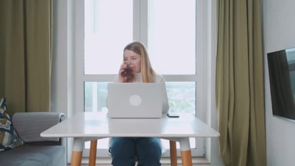Νεαρή γυναίκα Ευρωπαίος ελεύθερος επαγγελματίας εργάζεται στο σπίτι, στο τραπέζι χρησιμοποιώντας φορητό υπολογιστή, ενώ έχοντας ένα φλιτζάνι τσάι. — Αρχείο Βίντεο
