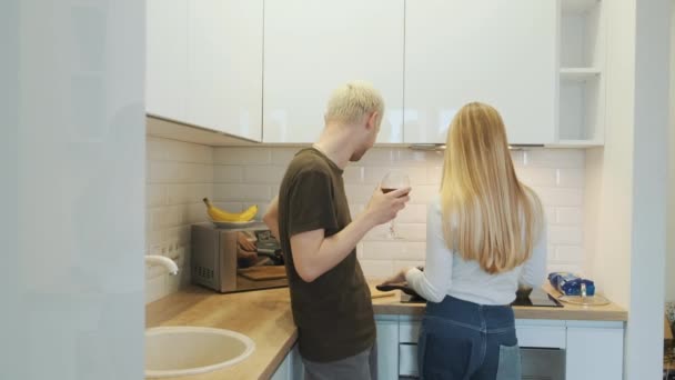 Joven feliz rubia europea pareja de amigos teniendo una conversación en la cocina moderna blanca mientras se cocina la cena. — Vídeo de stock
