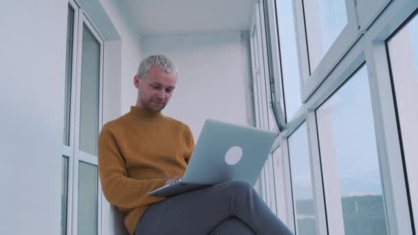 Portret van een jonge Europese mannelijke freelancer zittend op het balkon met de laptop multitasking. — Stockvideo