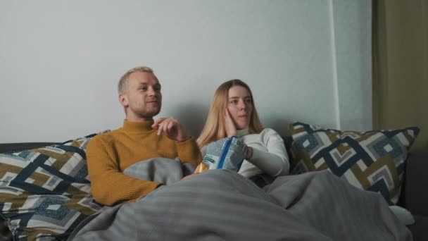 Jeune beau couple européen regarde un film à la maison. Assis sur le canapé sous la couverture et se nourrissant mutuellement puces. — Video