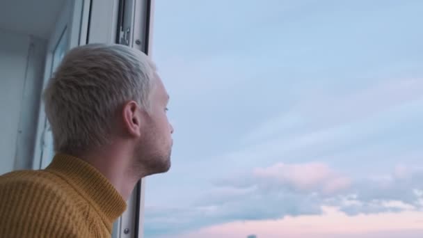 Primer plano de un tipo mirando por la ventana del balcón disfrutando de la vista del atardecer y mirando hacia el suelo. — Vídeos de Stock