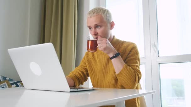 Mladý samec Evropan na volné noze pracuje doma, u stolu pomocí notebooku, zatímco si dává šálek čaje. — Stock video