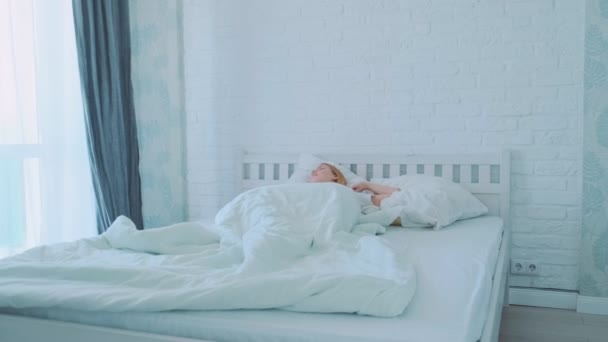 Όμορφη νεαρή Ευρωπαία κοπέλα ξυπνάει το πρωί και κοιτάζει το παράθυρο ενώ μένει στο κρεβάτι χασμουριέται. Λευκό μοντέρνο εσωτερικό. — Αρχείο Βίντεο