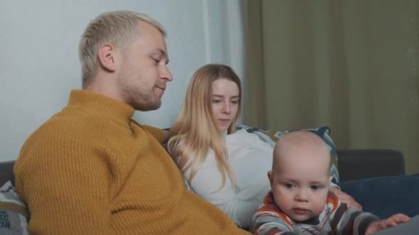 Unge smukke europæiske familie på tre ser en film derhjemme. Sidder på sofaen under tæppet og fodrer hinanden chips. – Stock-video