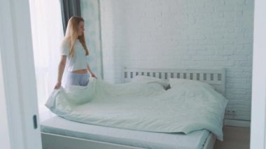 Güzel bir Avrupalı kızın, sabah pijama giyerek uyandıktan sonra yatağı toplarken yavaş çekimde. Beyaz ve modern iç mekan