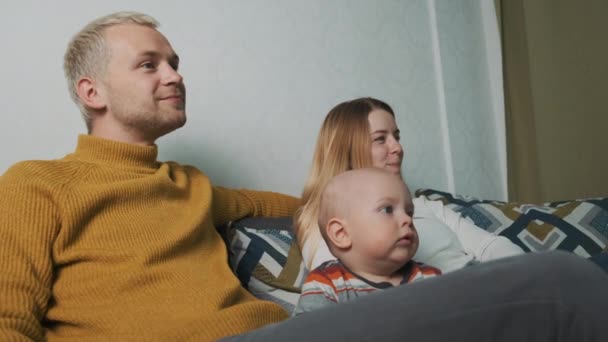 Mladá krásná evropská trojčlenná rodina se doma dívá na film. Sedět na gauči pod přikrývkou a krmit se hranolkama. — Stock video