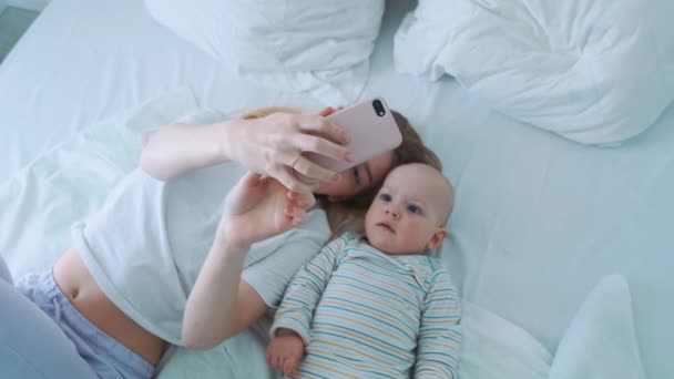 Όμορφο αγοράκι και η νεαρή μητέρα του που έχει ένα videocall ή τη λήψη selfies ενώ στο κρεβάτι το πρωί. Ευτυχισμένη οικογένεια. — Αρχείο Βίντεο
