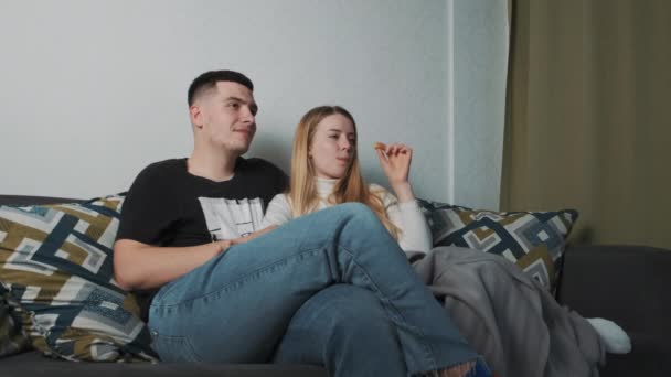 Mladý krásný evropský pár se doma dívá na film. Sedět na gauči pod přikrývkou a krmit se hranolkama. — Stock video
