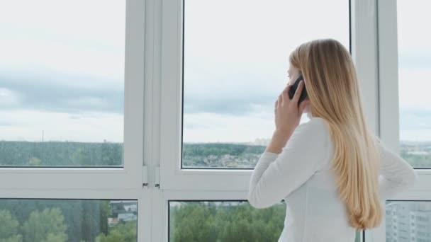 Junge energische Geschäftsfrau telefoniert mit einem Kunden vor einem riesigen Panoramafenster mit Blick auf die Stadt. — Stockvideo