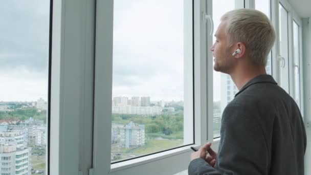 Νεαρός ενεργητικός επιχειρηματίας που μιλάει σε έναν πελάτη τηλεφωνικά μπροστά από ένα τεράστιο πανοραμικό παράθυρο με θέα στην πόλη. — Αρχείο Βίντεο