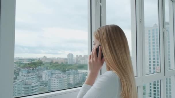 Junge energische Geschäftsfrau telefoniert mit einem Kunden vor einem riesigen Panoramafenster mit Blick auf die Stadt. — Stockvideo