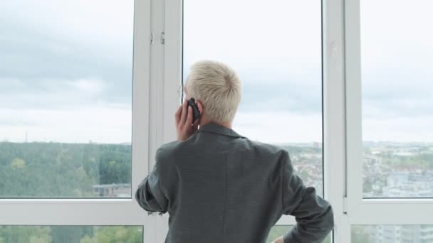 Joven empresario enérgico hablando con un cliente por teléfono frente a una enorme ventana panorámica con vista a la ciudad. — Vídeos de Stock