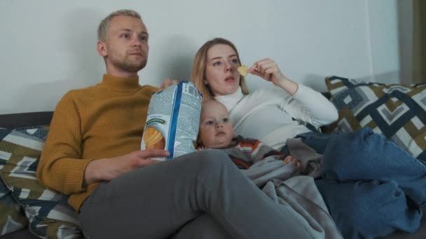 Молодая красивая европейская семья из трех человек смотрит кино дома. Сидя на диване под одеялом и кормя друг друга чипсами. — стоковое видео