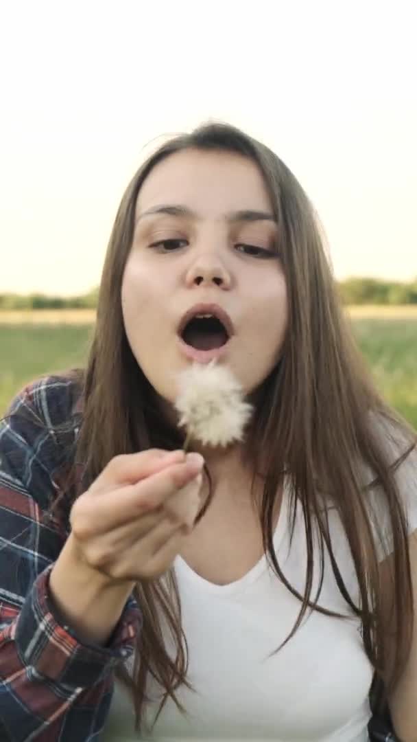 Ein junges Mädchen bläst einen Löwenzahn ab, den sie auf einem Feld gepflückt hat. Vertikales Video. — Stockvideo
