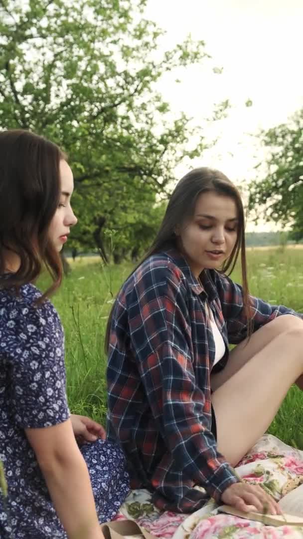 Zwei Schwestern wandeln in der Natur, kommunizieren, teilen Geheimnisse und lesen ein Buch. Vertikales Video. — Stockvideo