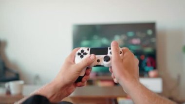 Geniş ekran bir televizyonun önündeki oyun konsolunda video oyunu oynayan bir gencin ellerini yakın plan çekiyoruz.